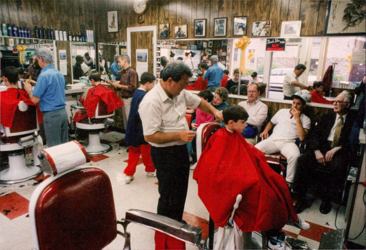 Barbers cutting customers’ hair inside Antonio’s Barber Shop, with waiting customers and photographs on the walls